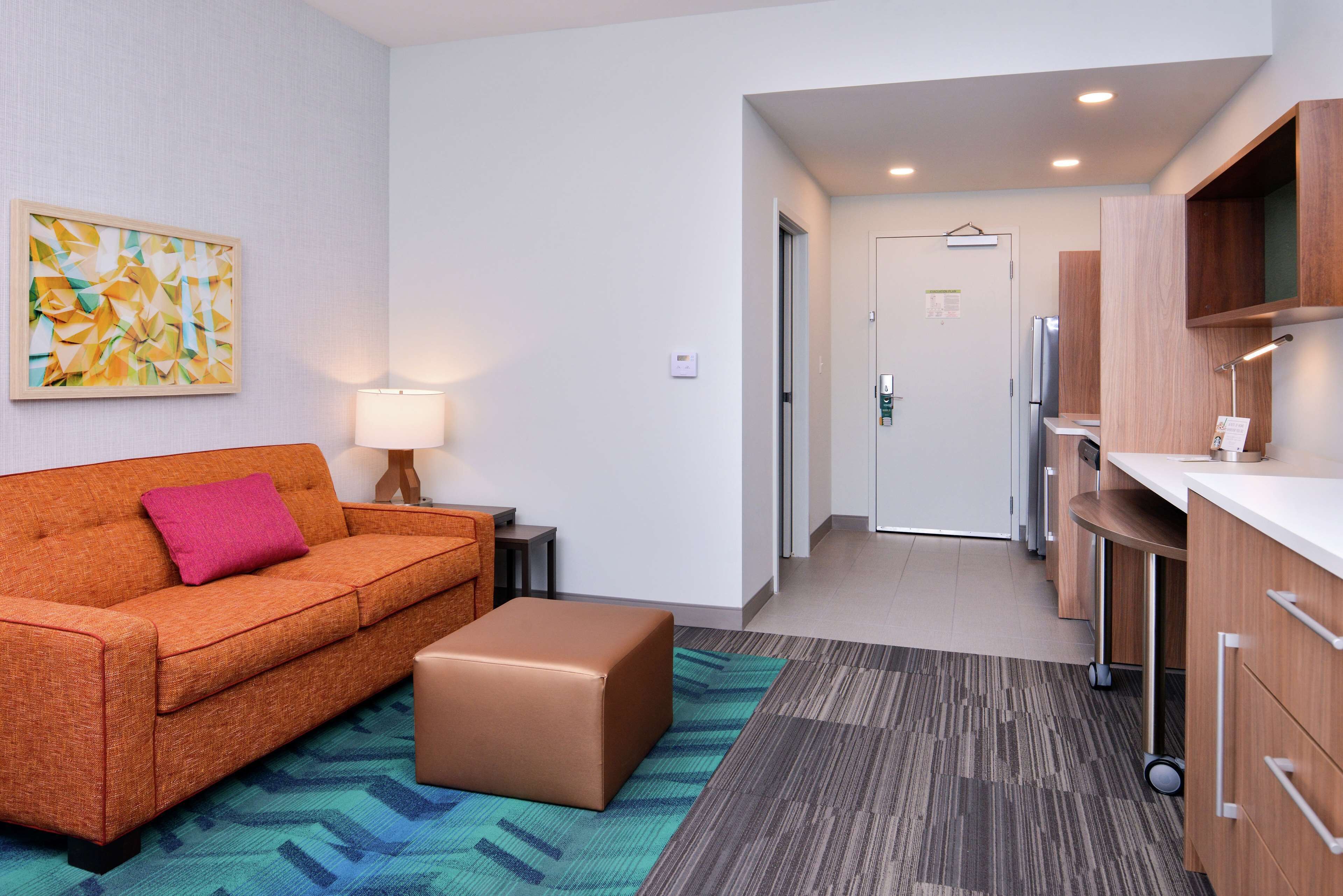 Una habitación de hotel moderna con sofá, un pequeño taburete, lámpara y zona de cocina con armarios y refrigerador.