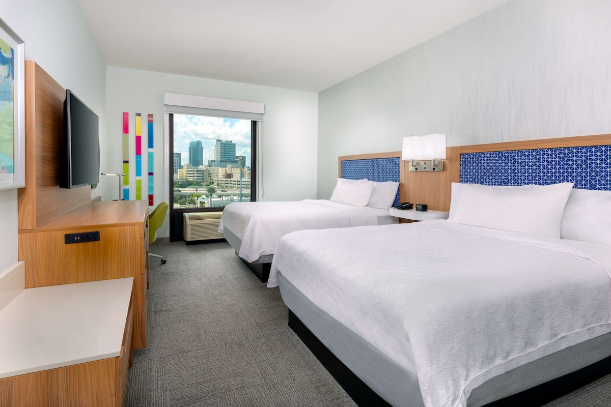 A hotel room with two beds, a desk, a TV, and a city view.