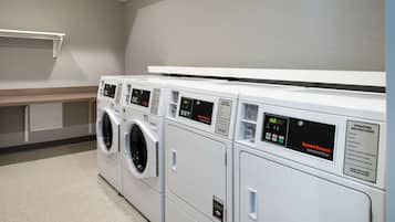 A row of white commercial dryers with control panels, a white shelf above, and a grey wall.