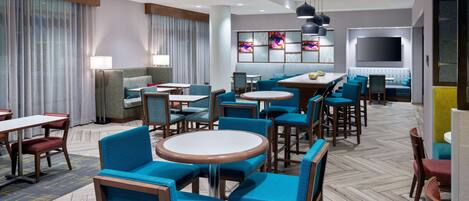 Um hall de hotel com várias áreas de estar, incluindo mesas redondas e cadeiras, um sofá e uma televisão na parede.