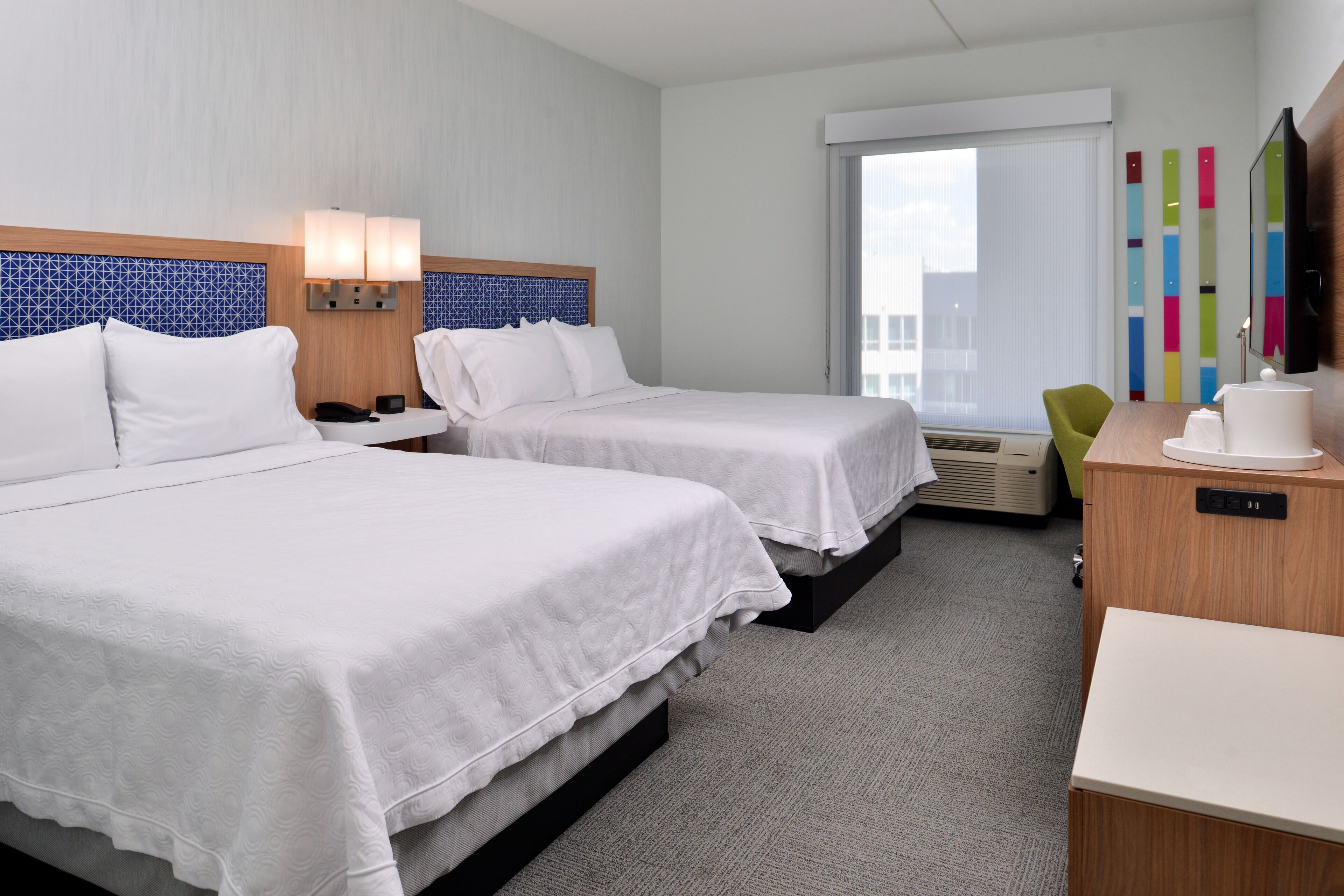 A hotel room with two beds, a desk, a chair, a TV, and a window with blinds.