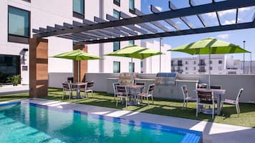 Un área de piscina en la azotea con una zona de barbacoa, asientos exteriores y sombrillas.
