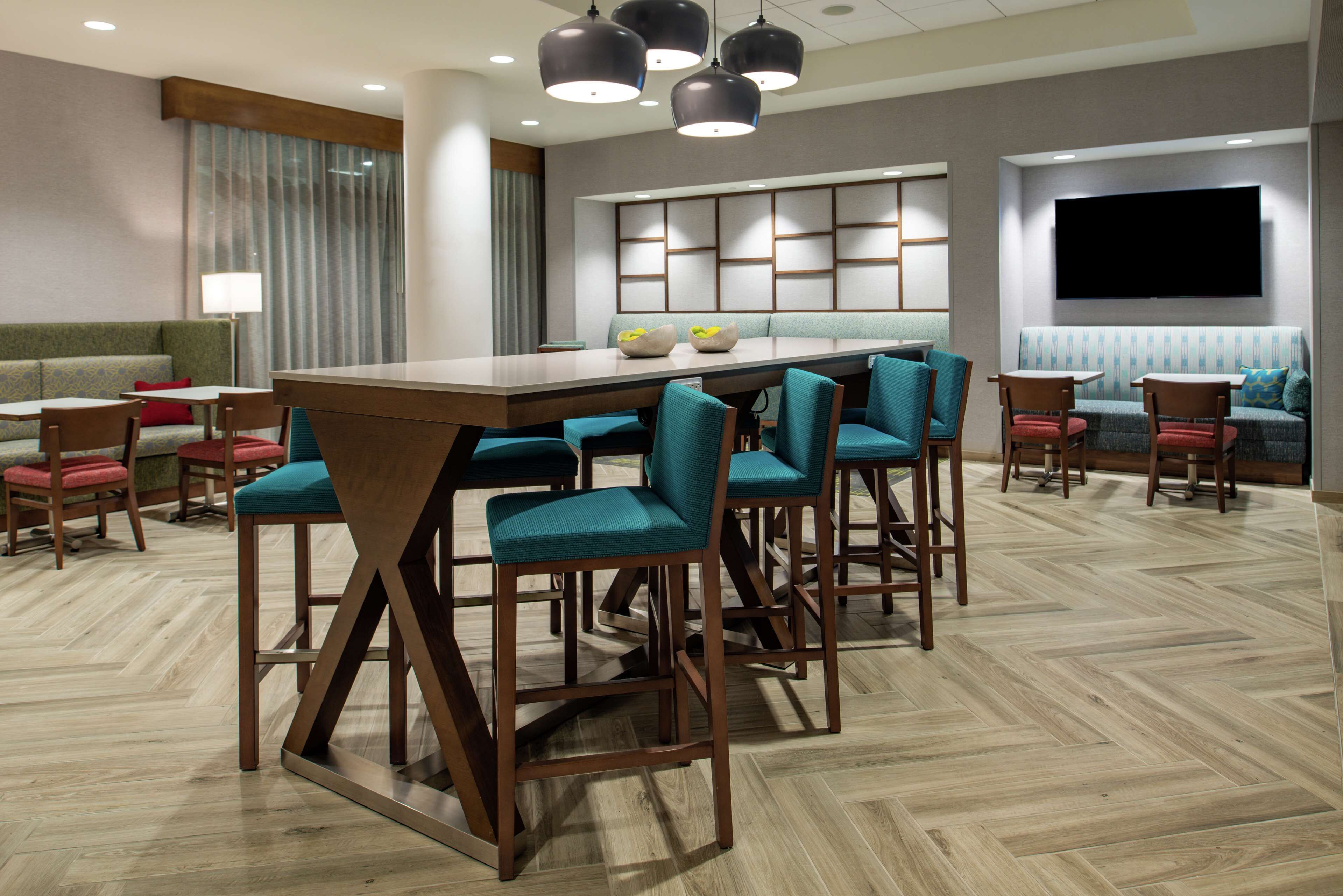 A modern hotel lobby with a long dining table, multiple bar stools, a sofa, and a TV mounted on the wall.