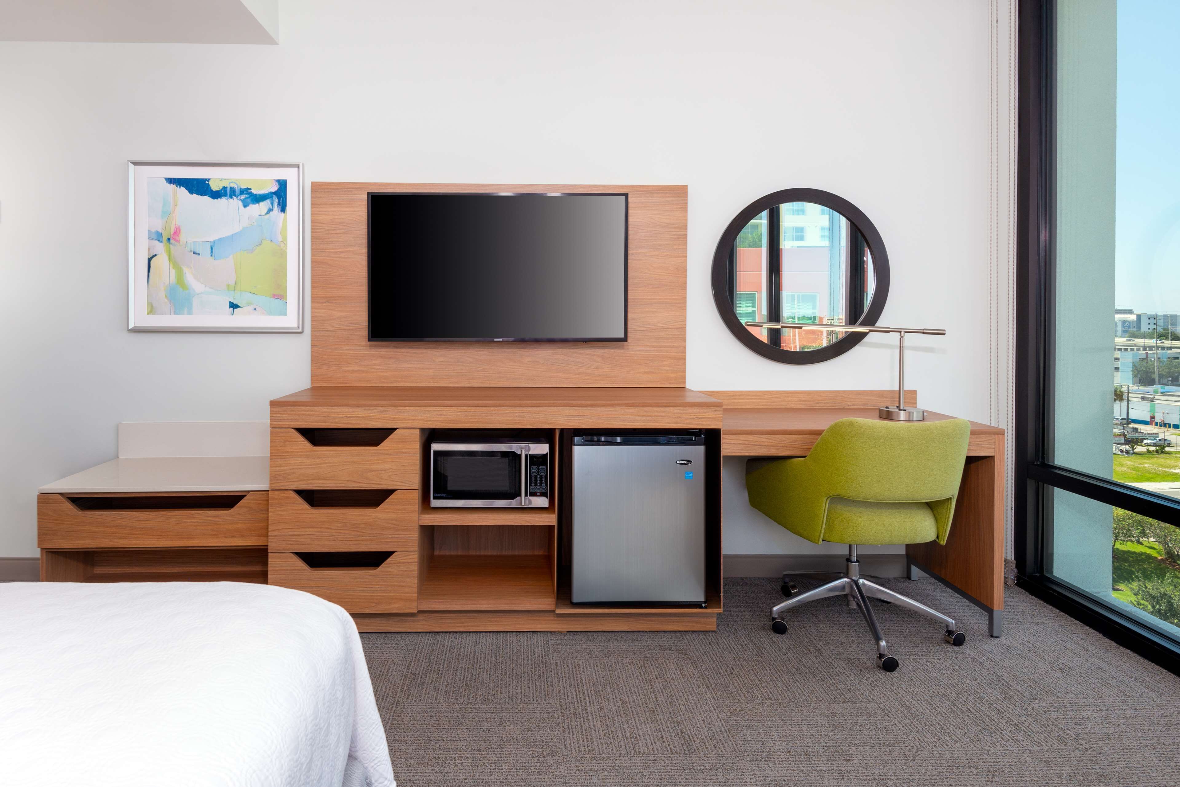A hotel room with a bed, a desk with a flat-screen TV, a microwave, a mini-fridge, a green chair, and a large window offering a city view.