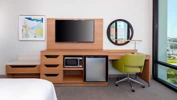 A hotel room with a bed, a desk with a flat-screen TV, a microwave, a mini-fridge, a green chair, and a large window offering a city view.