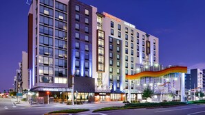 Un hôtel à plusieurs étages, reconnaissable à son grand panneau « Hampton Inn », à son café « Starbucks » et à son entrée moderne agrémentée d’un auvent coloré.