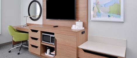 Una habitación de hotel moderna con un escritorio de madera, un televisor de pantalla plana, un microondas, una silla verde y un cuadro abstracto enmarcado.