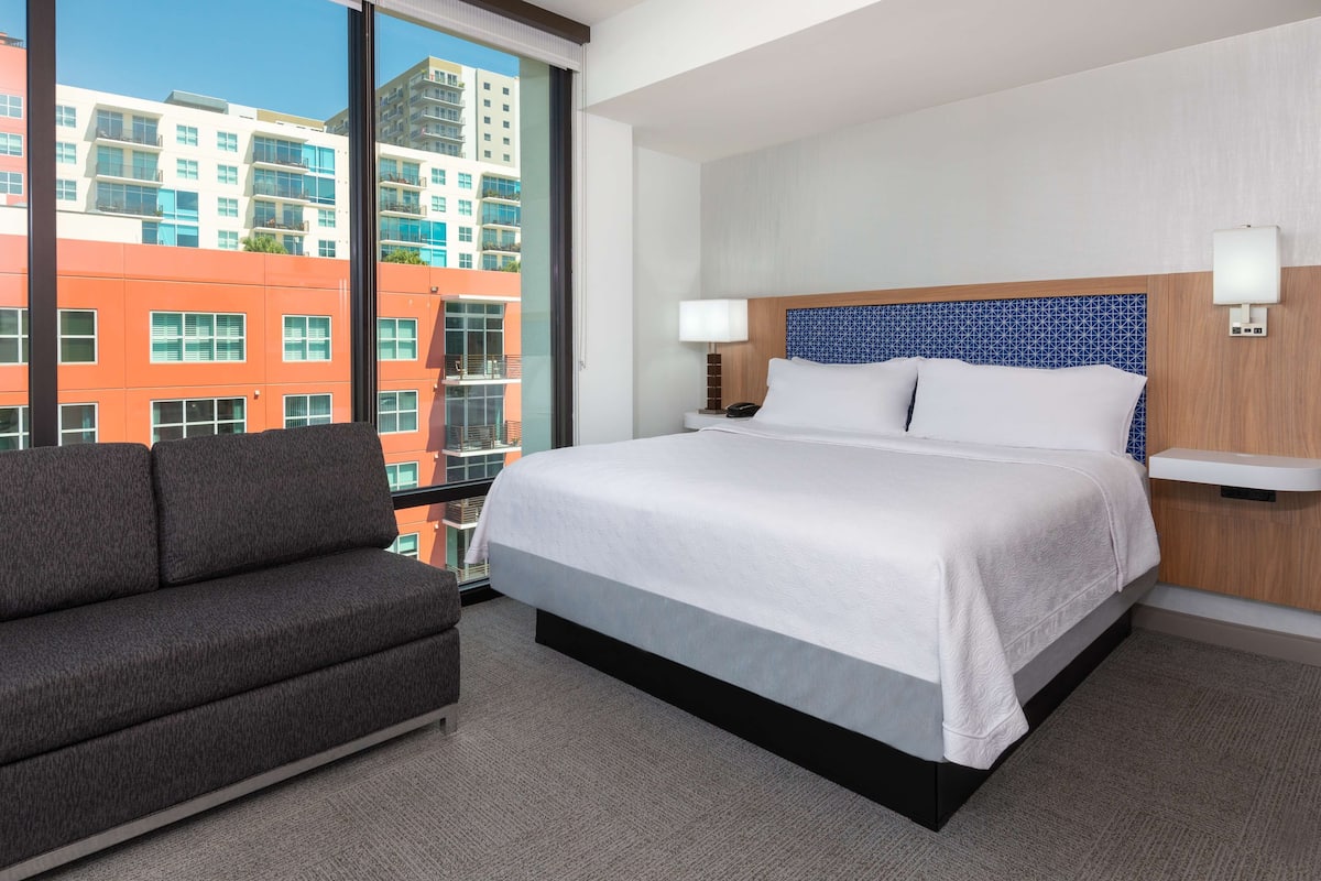 A hotel room with a large bed, a sofa, a nightstand, and a view of buildings through a large window.