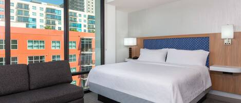 A hotel room with a large bed, a sofa, a nightstand, and a view of buildings through a large window.