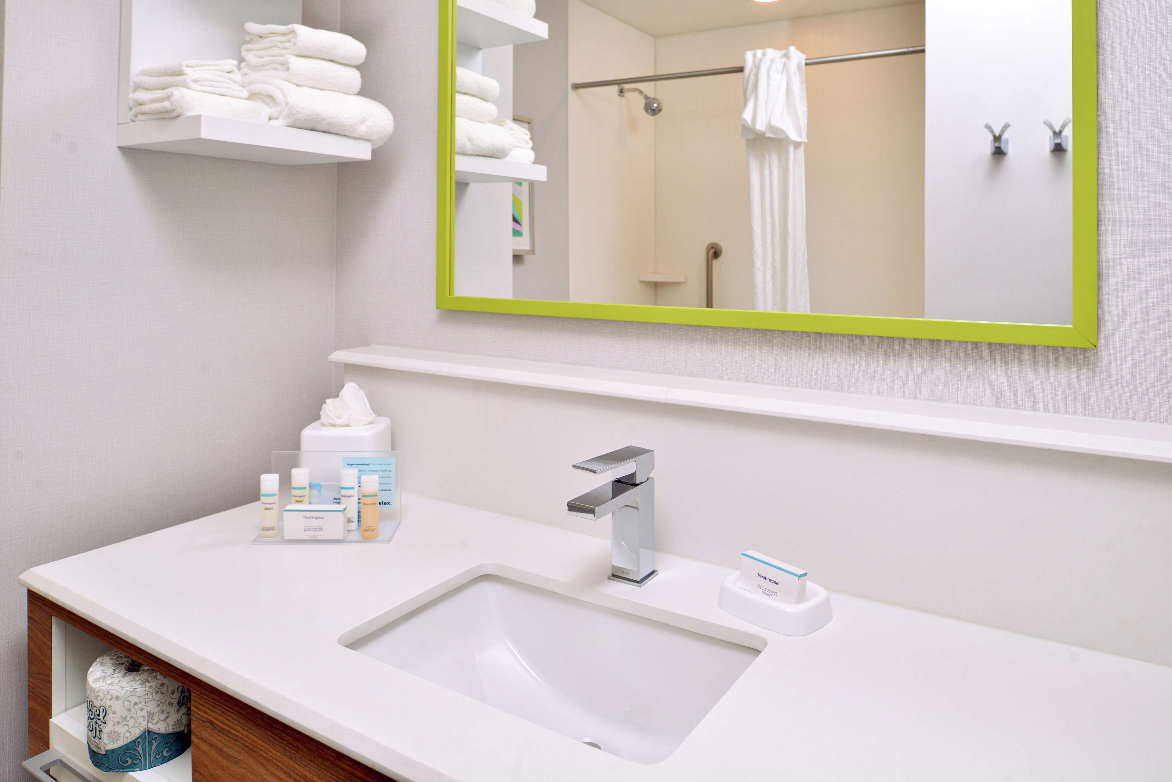 A modern bathroom with a white sink, a green-framed mirror, and neatly folded towels on a shelf.