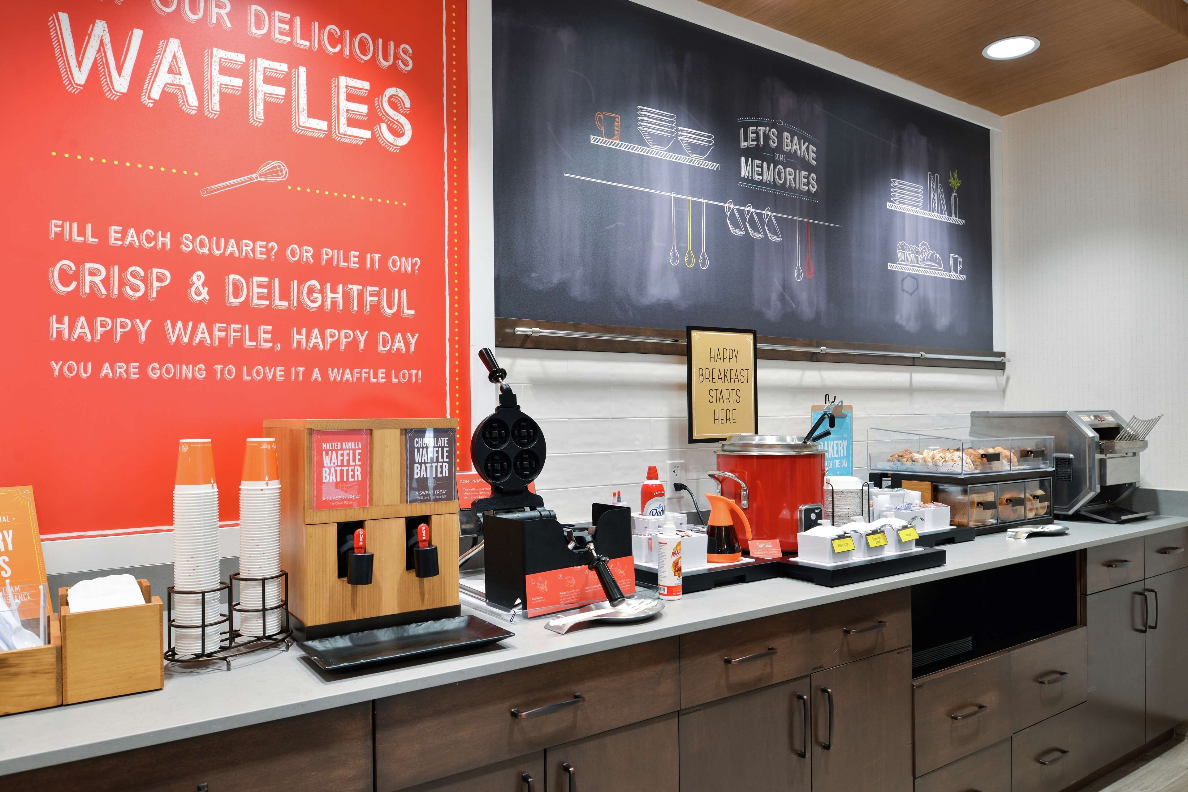 Una estación de waffles con un menú en una pizarra, dispensadores de masa para gofres y una máquina de café.