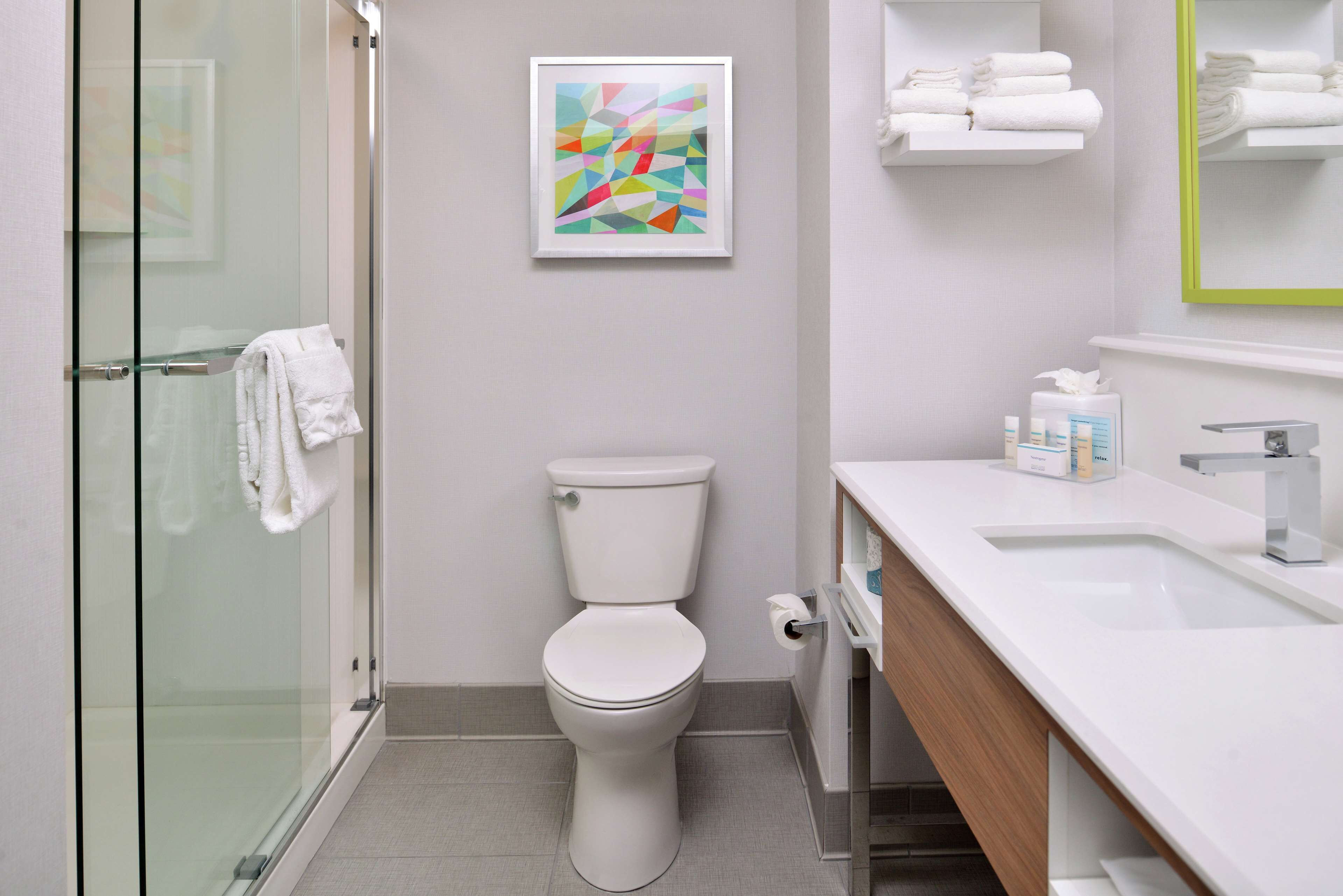 A modern bathroom with a glass shower enclosure, a toilet, a sink with a mirror, and a framed abstract painting on the wall.