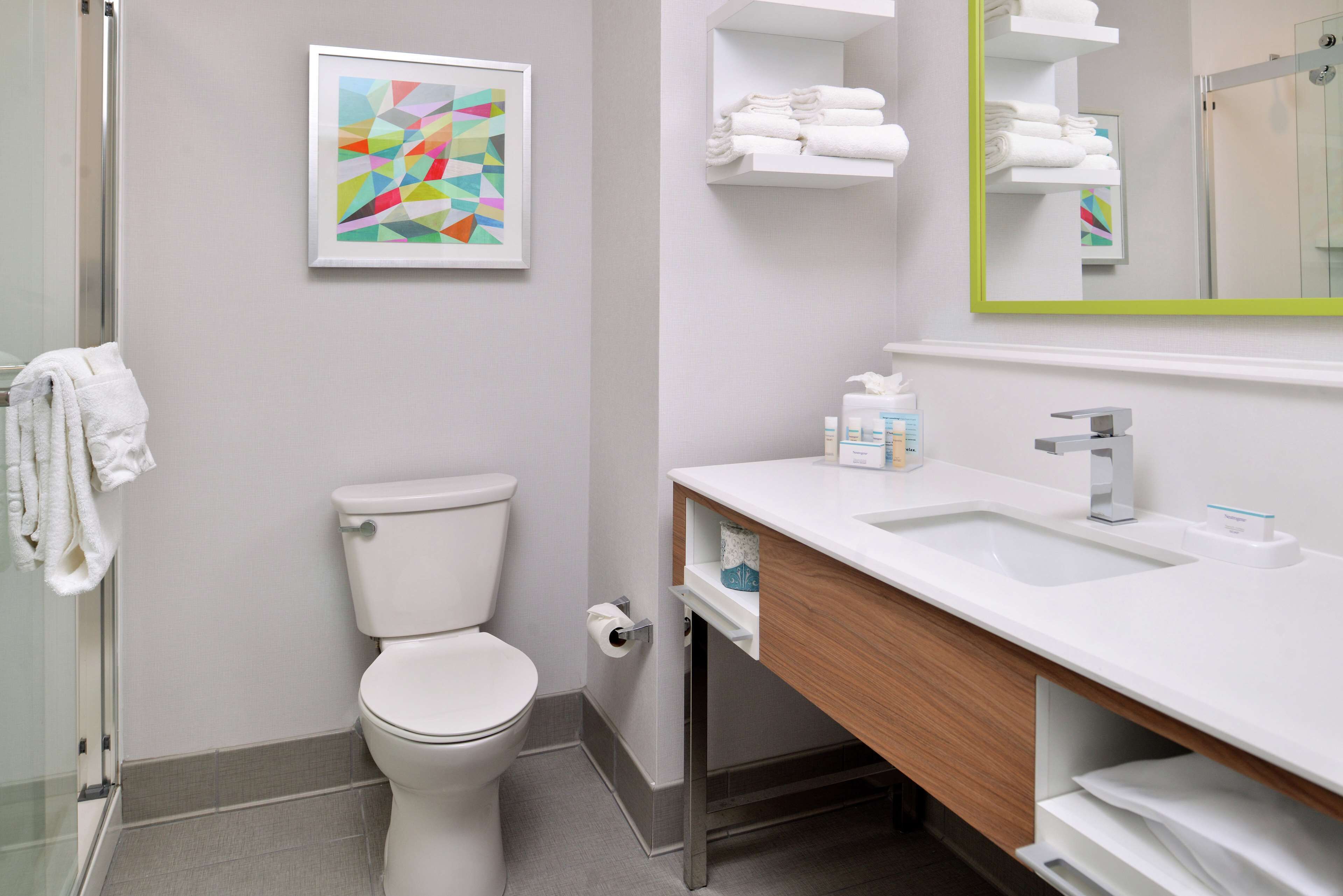 Ein modernes Badezimmer mit Toilette, Waschbecken und Dusche. An der Wand hängt ein gerahmtes abstraktes Kunstwerk und ein Spiegel mit grünem Rahmen.