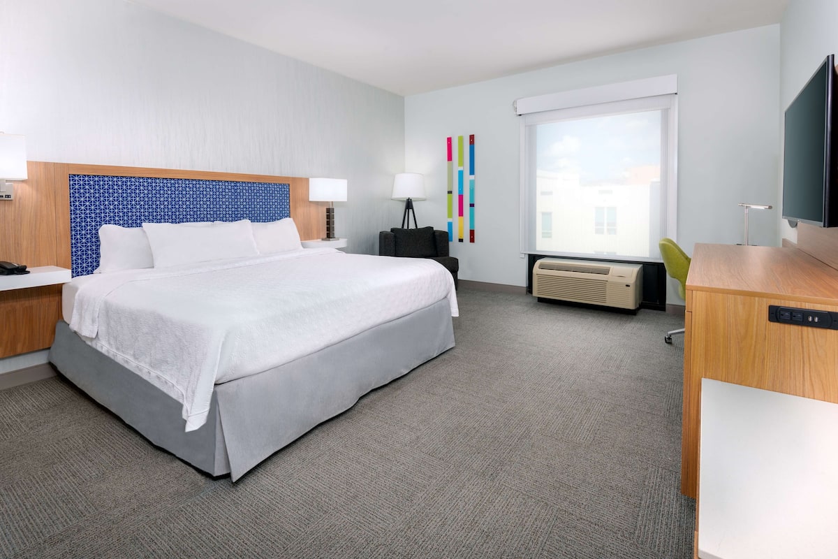 A hotel room with a large bed, a desk, a chair, a TV, and a window with a city view.