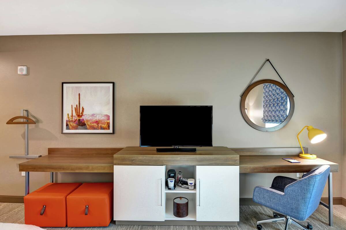 Ein modernes Hotelzimmer mit Schreibtisch aus Holz, einem Flachbildfernseher, einem runden Spiegel, einem gerahmten Bild, einer Schreibtischlampe, einem blauen Bürostuhl und Stauraum.