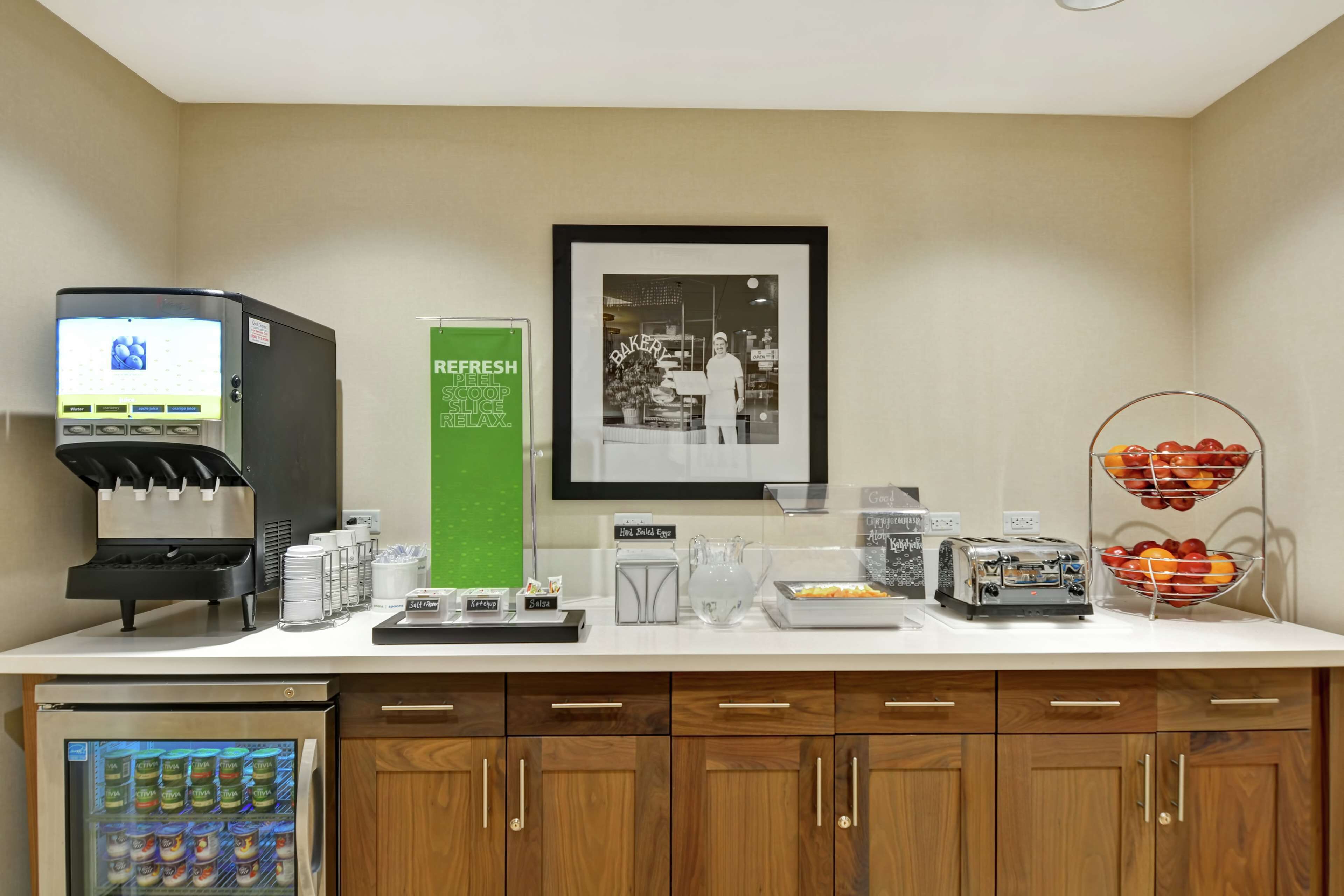 A kitchenette with a coffee machine, a refrigerator, a framed picture, and a fruit display.