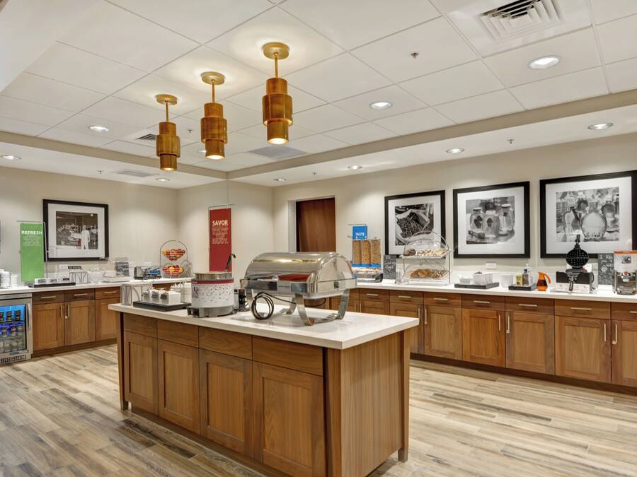 Un área de desayuno de hotel con una isla central, armarios de madera, una máquina de café y un dispensador refrigerado de bebidas.