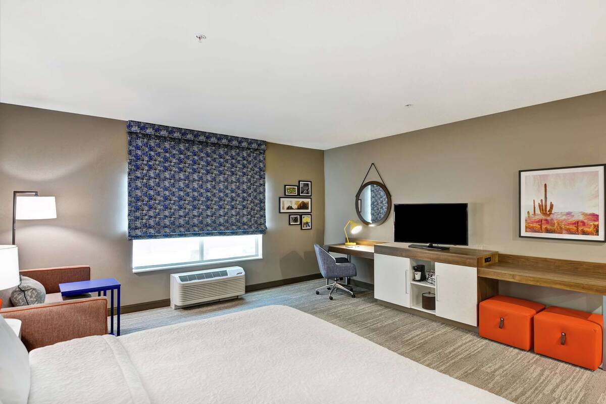 Ein Hotelzimmer mit einem Bett, einem Schreibtisch mit Computer, einem Fernseher und einem Fenster mit gemusterten Vorhängen.