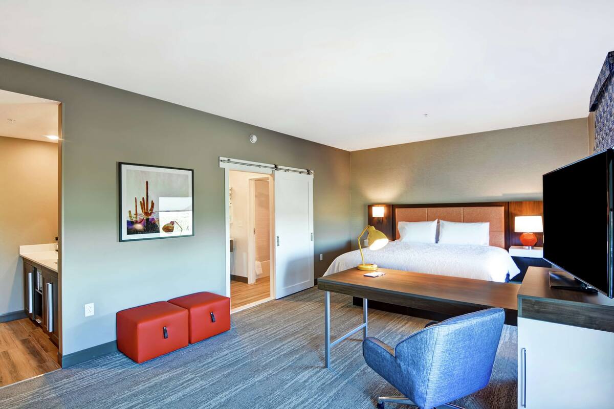 Ein Hotelzimmer mit Bett, Schreibtisch, Stuhl und einer roten Bank. An der Wand hängt ein Gemälde und auf dem Schreibtisch steht ein Fernseher.