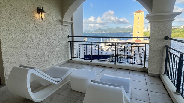 Un balcon avec des fauteuils de jardin blancs et une vue sur la mer et un bâtiment jaune.