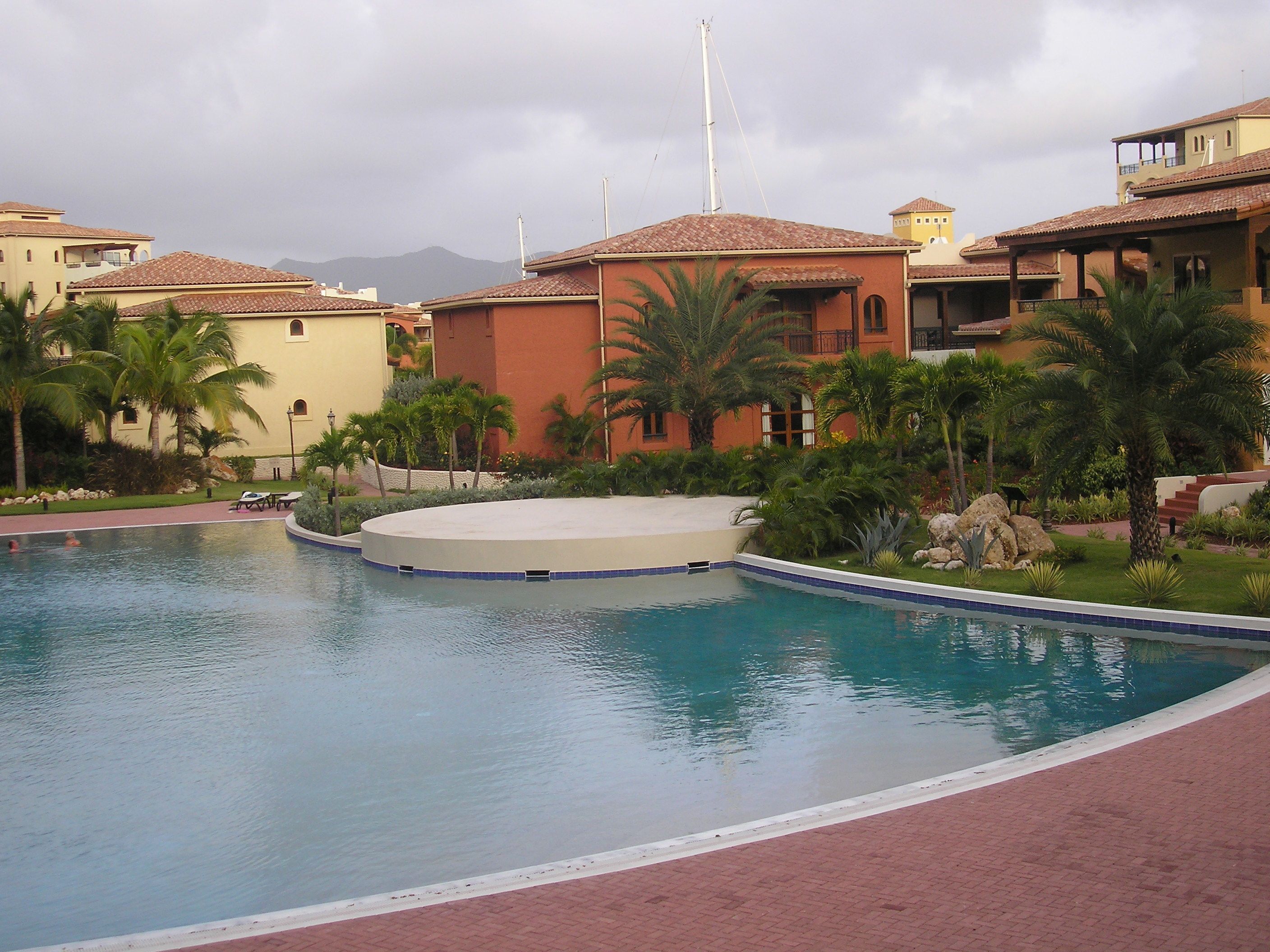 A swimming pool with a circular deck, surrounded by palm trees and buildings with red-tiled roofs.