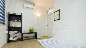 A room with a wooden floor, a white wall with a textured pattern, a black shelf with various items, a white door, and a small mirror on the wall. - Yours Guesthouse (Tongyeong)