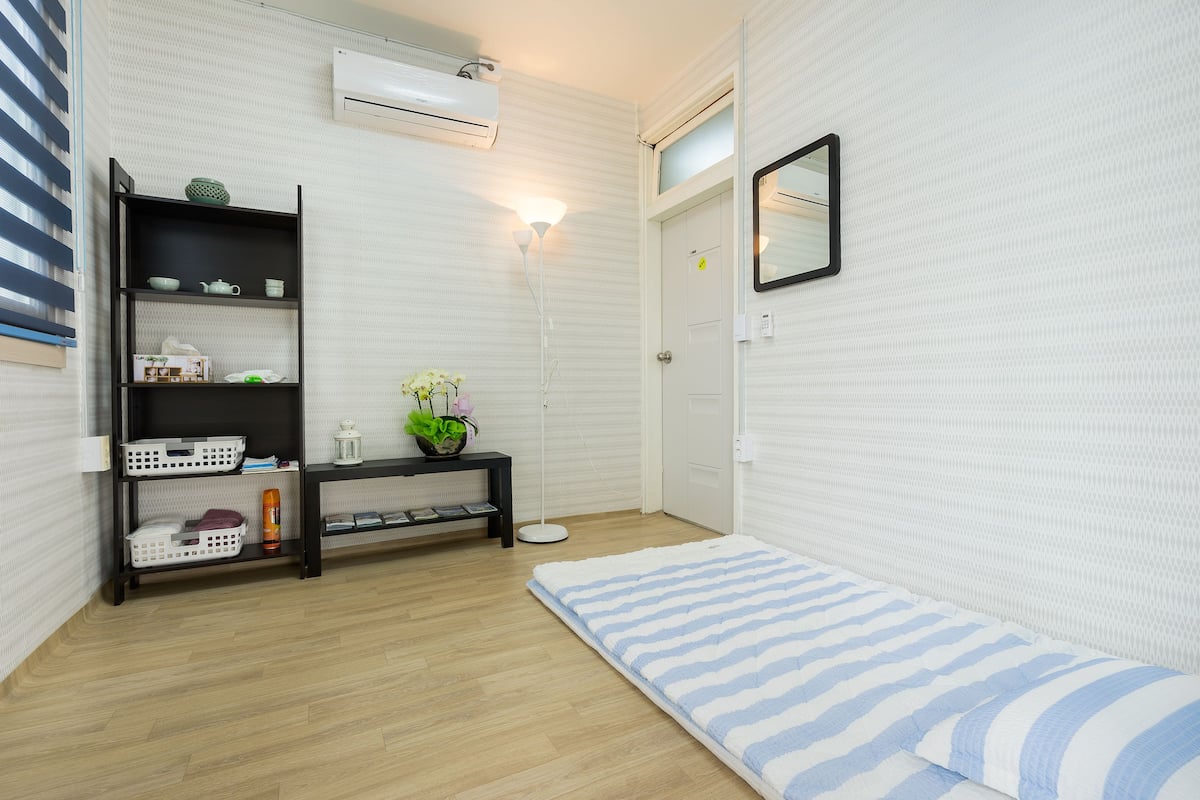 Ein Zimmer mit Holzboden, einer weißen Wand mit strukturierter Oberfläche, einem schwarzen Regal mit verschiedenen Gegenständen, einer weißen Tür und einem kleinen Spiegel an der Wand.