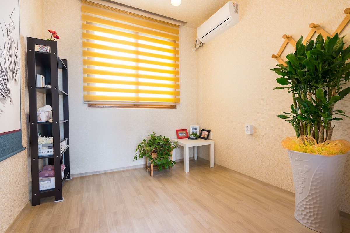 Ein Zimmer mit Holzboden, weißer Wand, einem Fenster mit Jalousien, einem weißen Tisch mit Pflanze, einem schwarzen Regalelement und einer weißen Vase mit Pflanze.