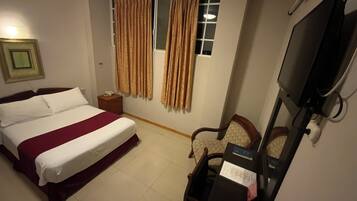 Habitación de hotel con una cama individual, un escritorio con silla, un televisor y cortinas en la ventana.