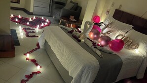 Una cama bien hecha con pétalos de rosa y velas, globos rosas y una mesita de noche con lámpara.
