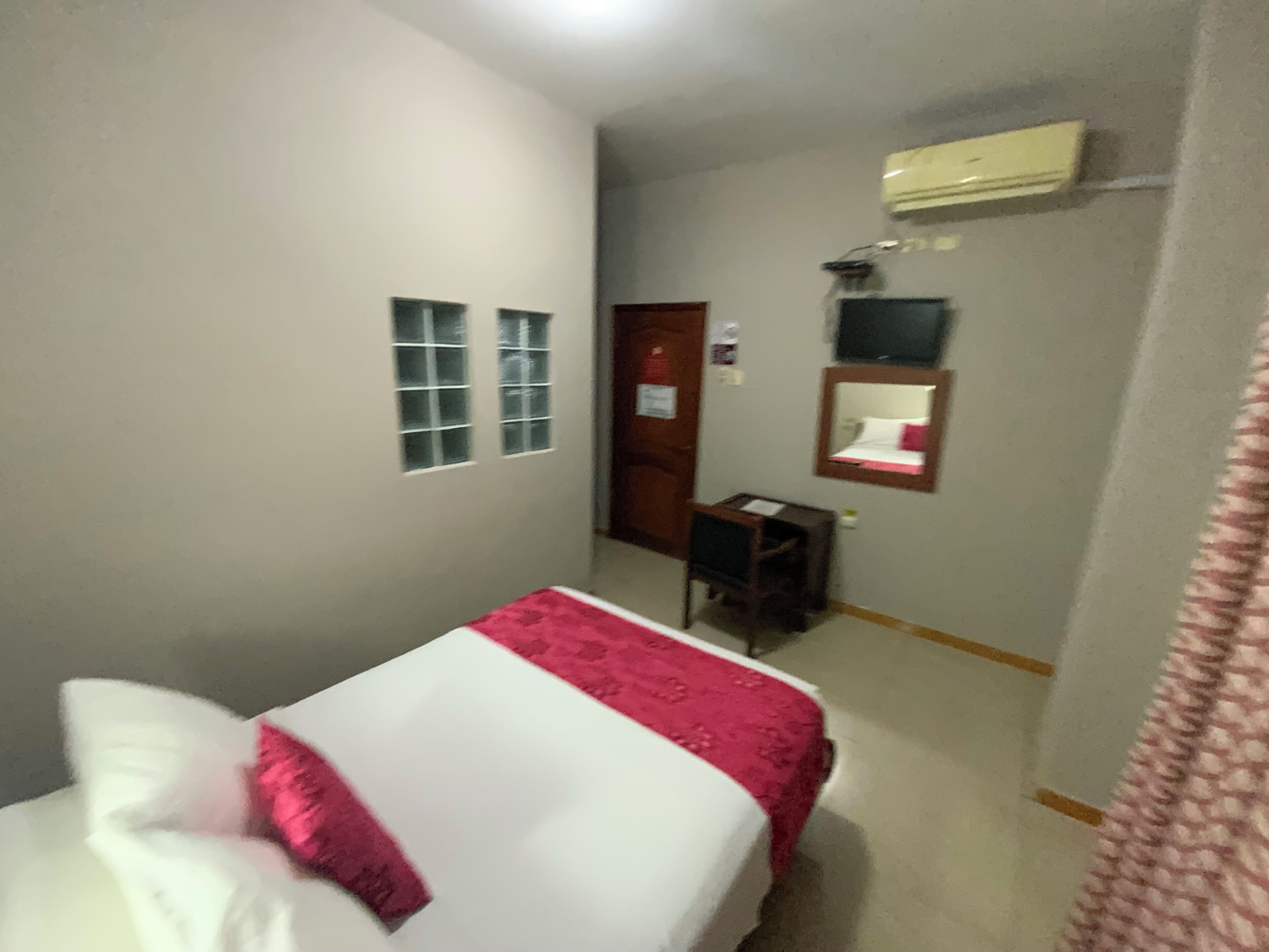 Quarto de hotel pequeno e estreito, com cama, escrivaninha, espelho e porta.