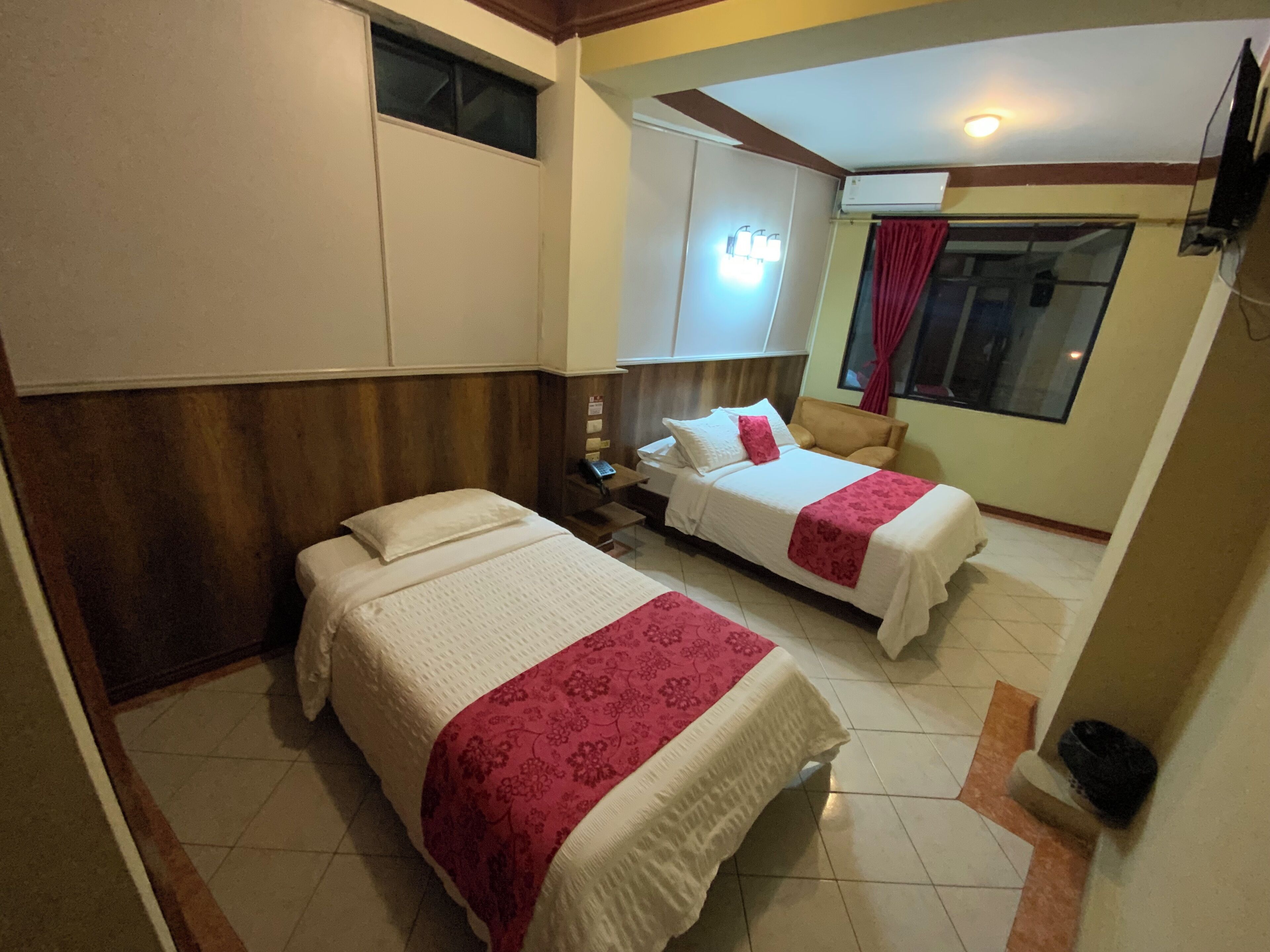 Ein Hotelzimmer mit zwei Betten, einem Nachttisch, einem kleinen Tisch, einem Fenster mit Vorhängen und einem Deckenventilator.