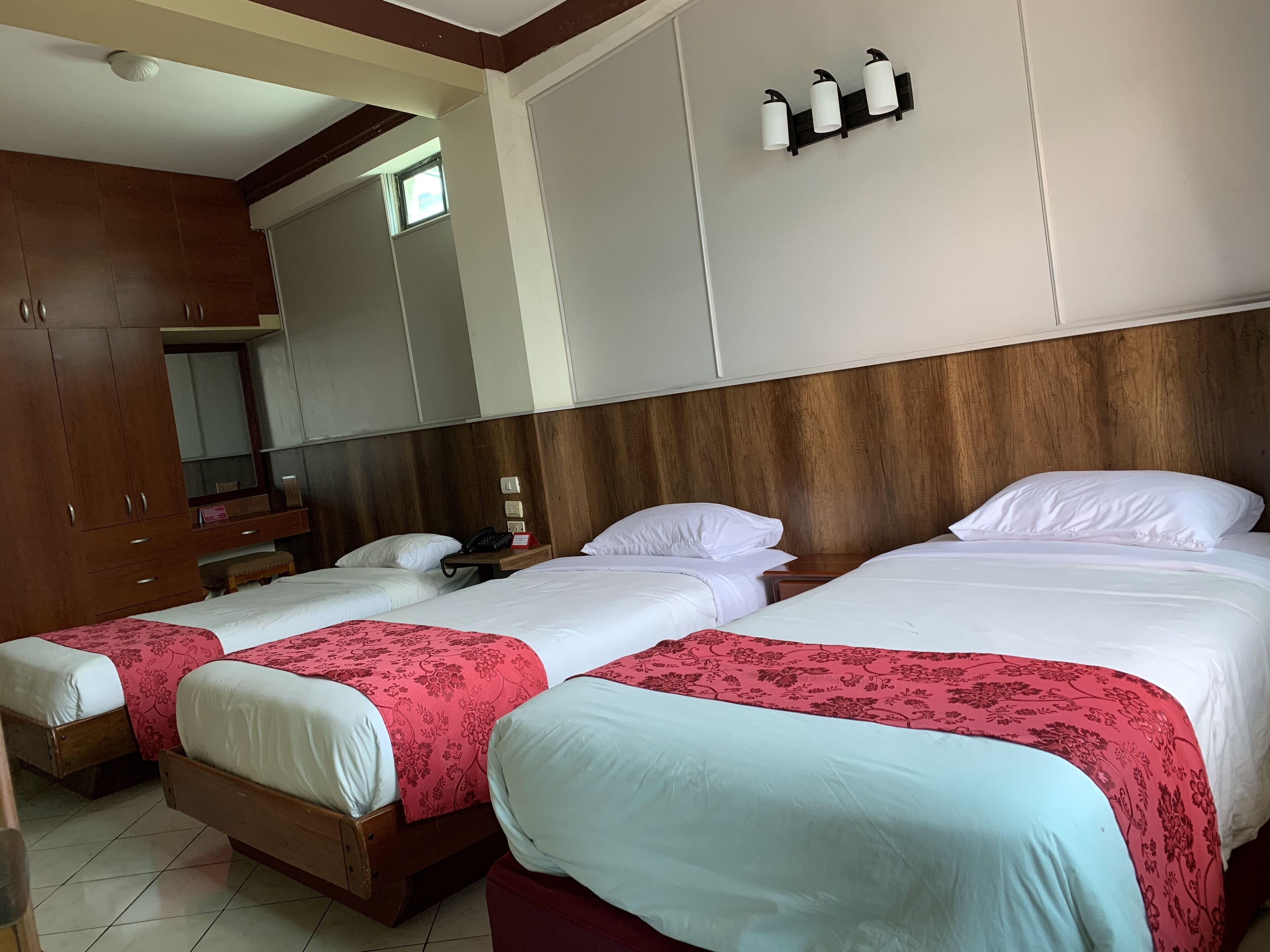 Habitación de hotel con dos camas individuales, un armario de madera, un escritorio pequeño con teléfono y una lámpara fijada a la pared.