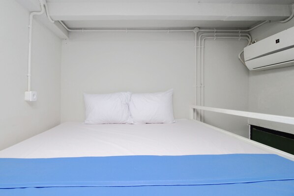 Una habitación pequeña con cama individual, paredes blancas, colcha azul y aire acondicionado blanco montado en la pared.