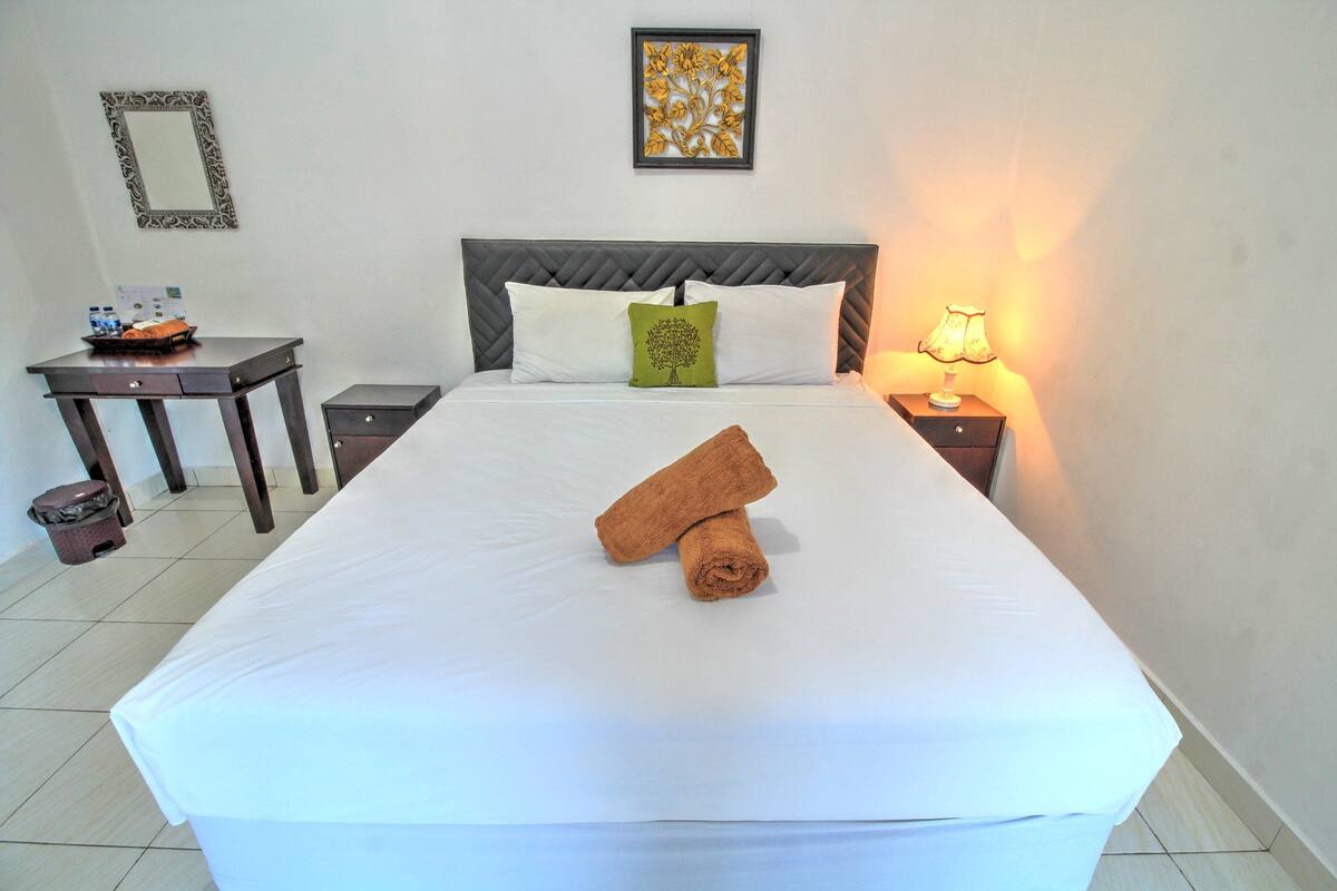 A neatly made bed with white linens, a green pillow, and two rolled towels. There is a bedside table with a lamp and a framed picture on the wall.