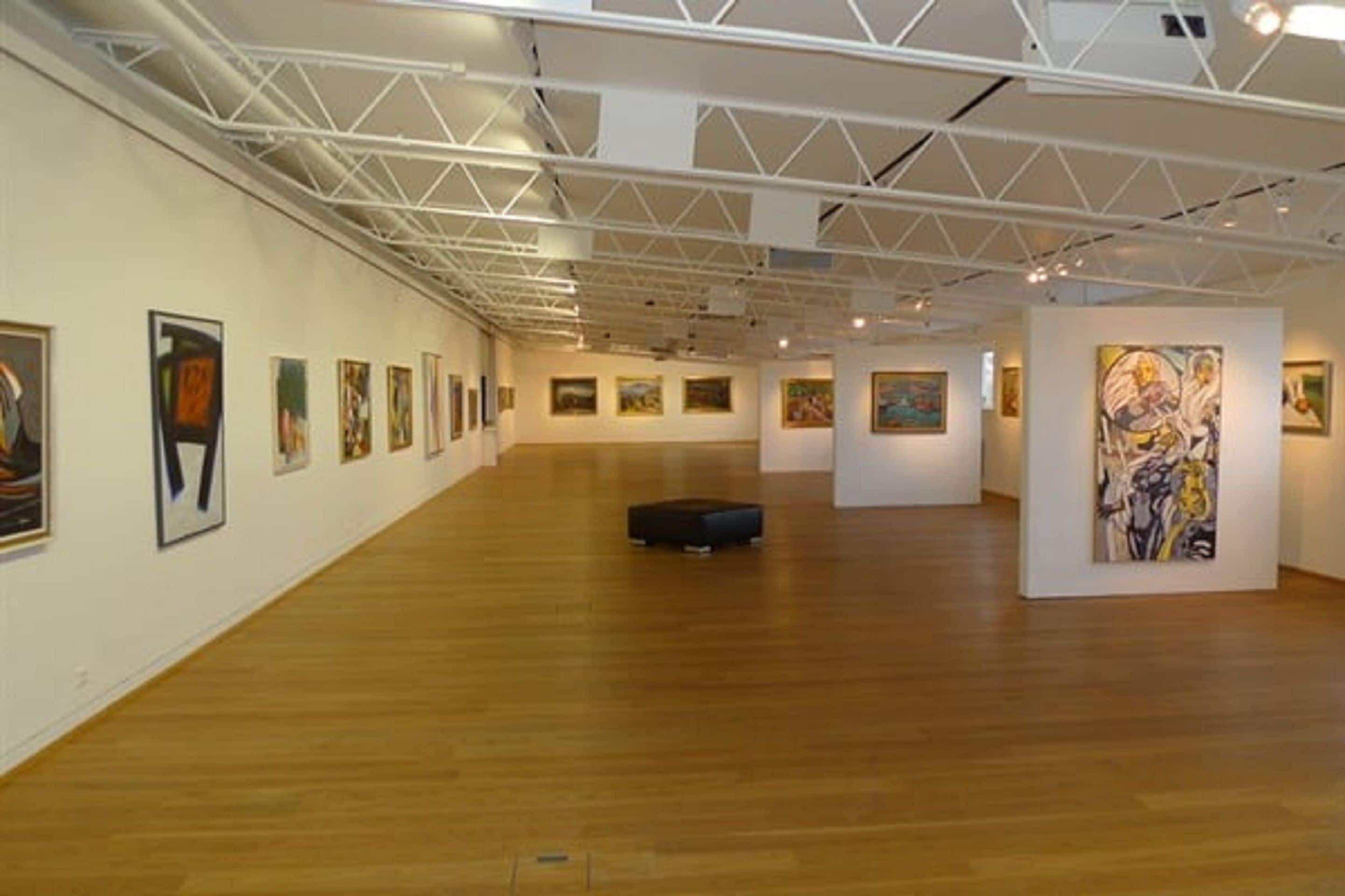 An art gallery with wooden floors and white walls, featuring several paintings and a black ottoman.