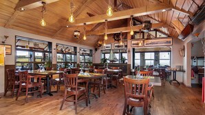 A restaurant with wooden floors, tables, and chairs, featuring large windows and hanging light fixtures. - Hotel Duus (Reykjanesbær)