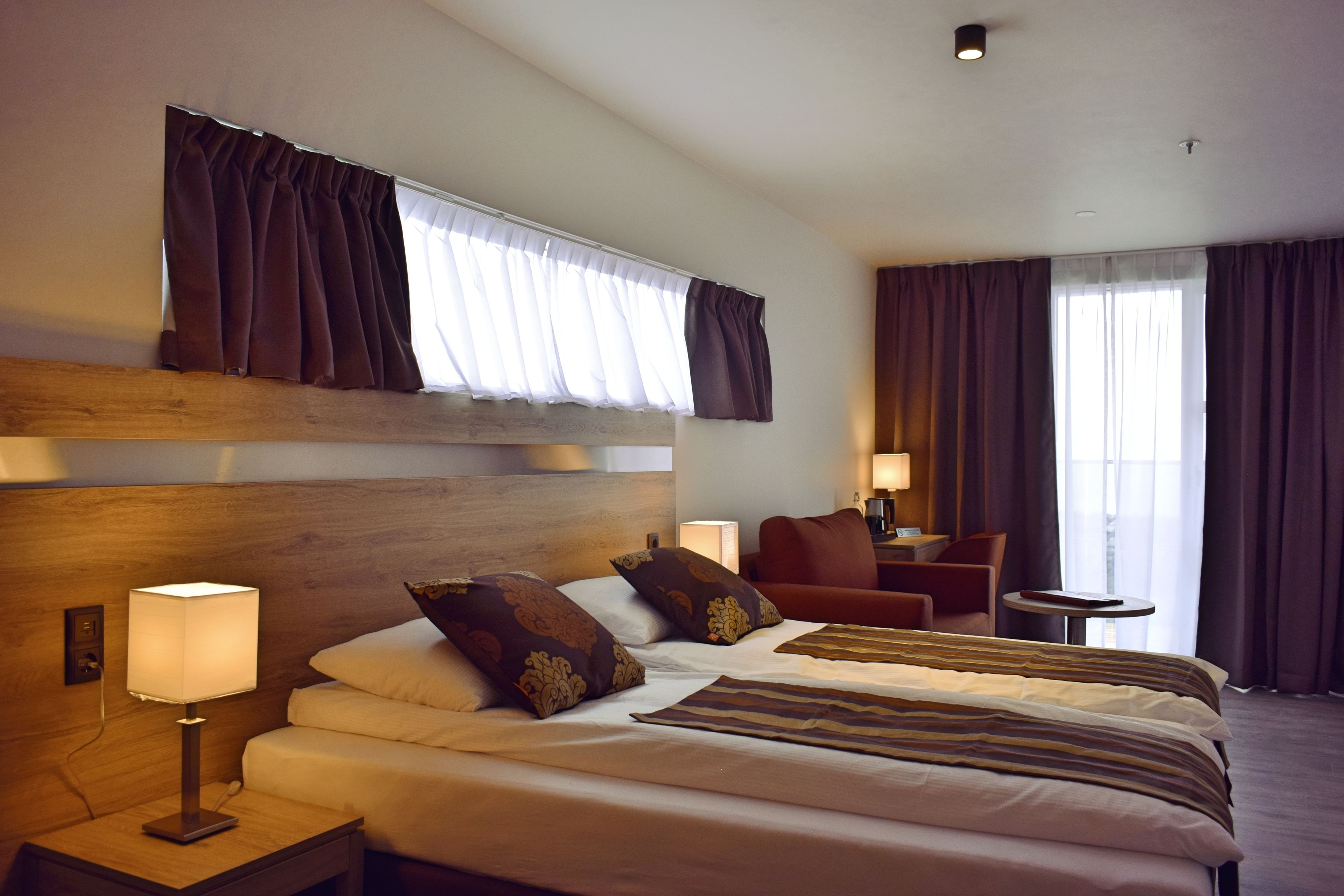 Ein Hotelzimmer mit einem großen Bett, einem Nachttisch mit Lampe, einem kleinen Tisch mit Stuhl und einem Fenster mit Vorhängen.