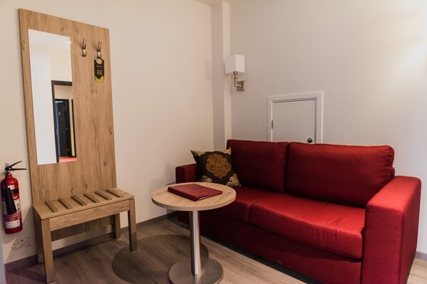 A room with a wooden bench, a red sofa, a round table with a book, a wall-mounted mirror, a fire extinguisher, and a door. - Hotel Duus (Reykjanesbær)