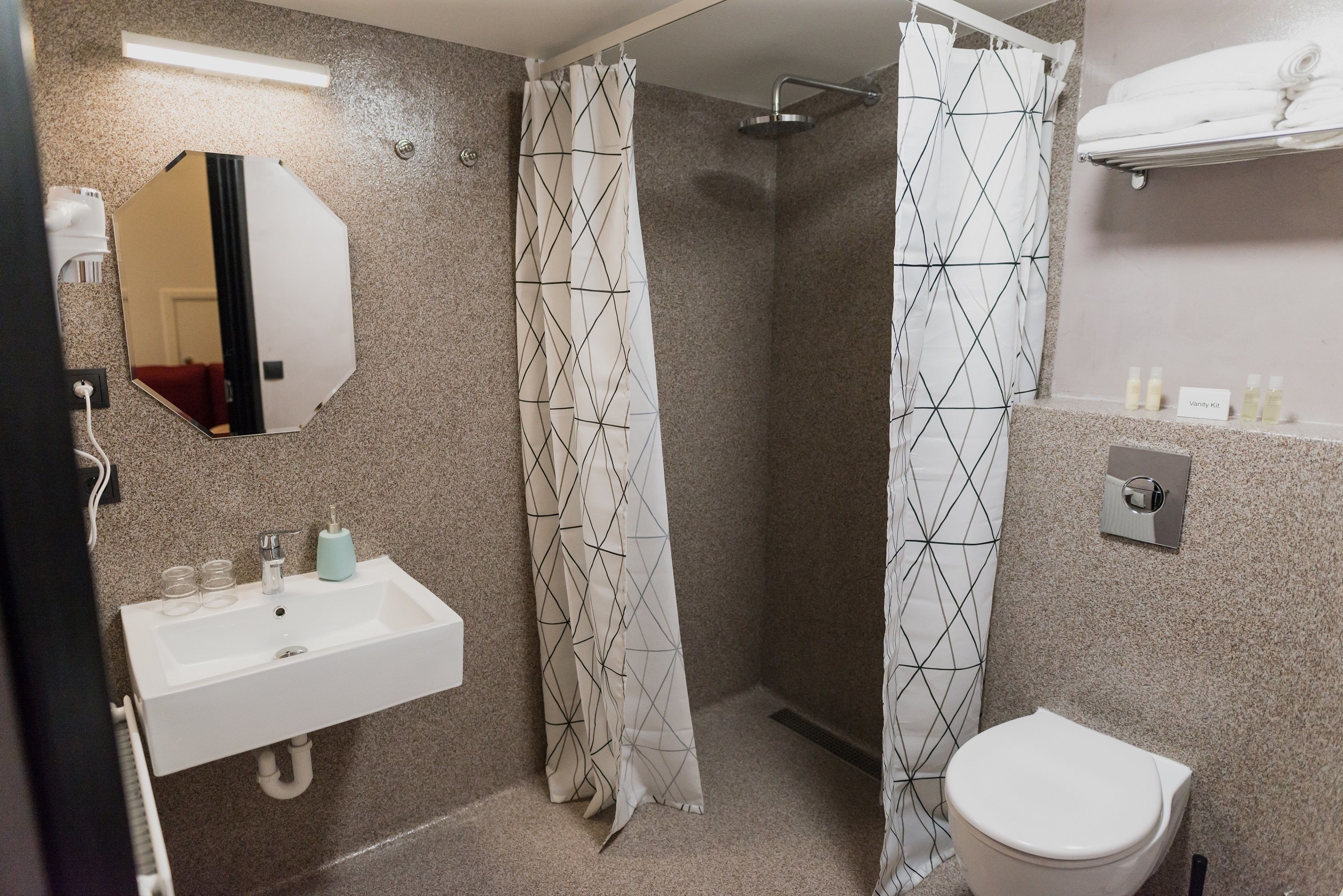 Un baño con lavamanos blanco, un espejo, una ducha con cortina de estampado geométrico y un inodoro.