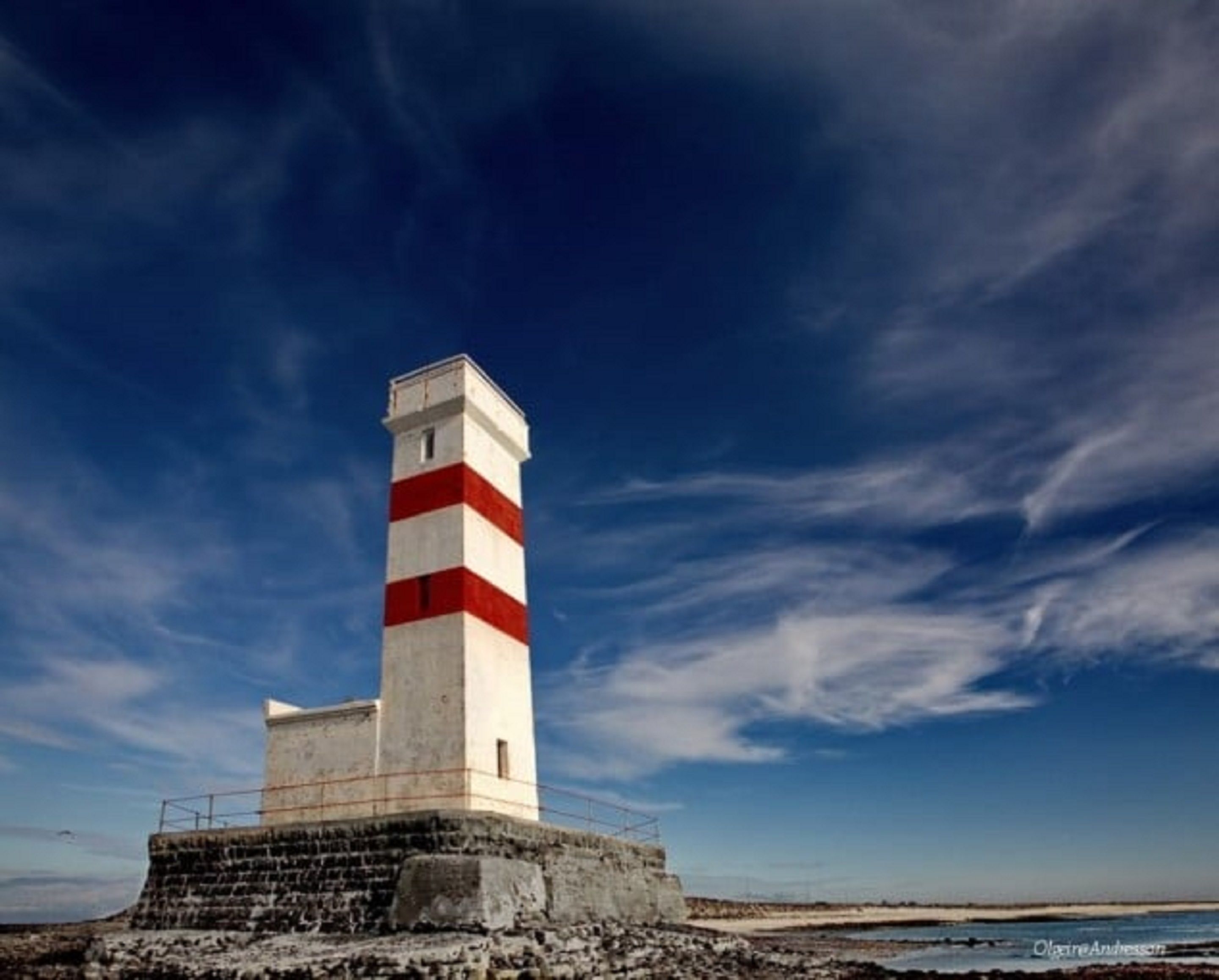 Un faro con una torre a rayas blancas y rojas, asentado sobre una base rocosa, con un cielo azul despejado de fondo.