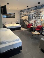 Una habitación de hotel moderna con un mural grande, dos camas, un área de comedor con sillas rojas y una pequeña mesa con un florero.