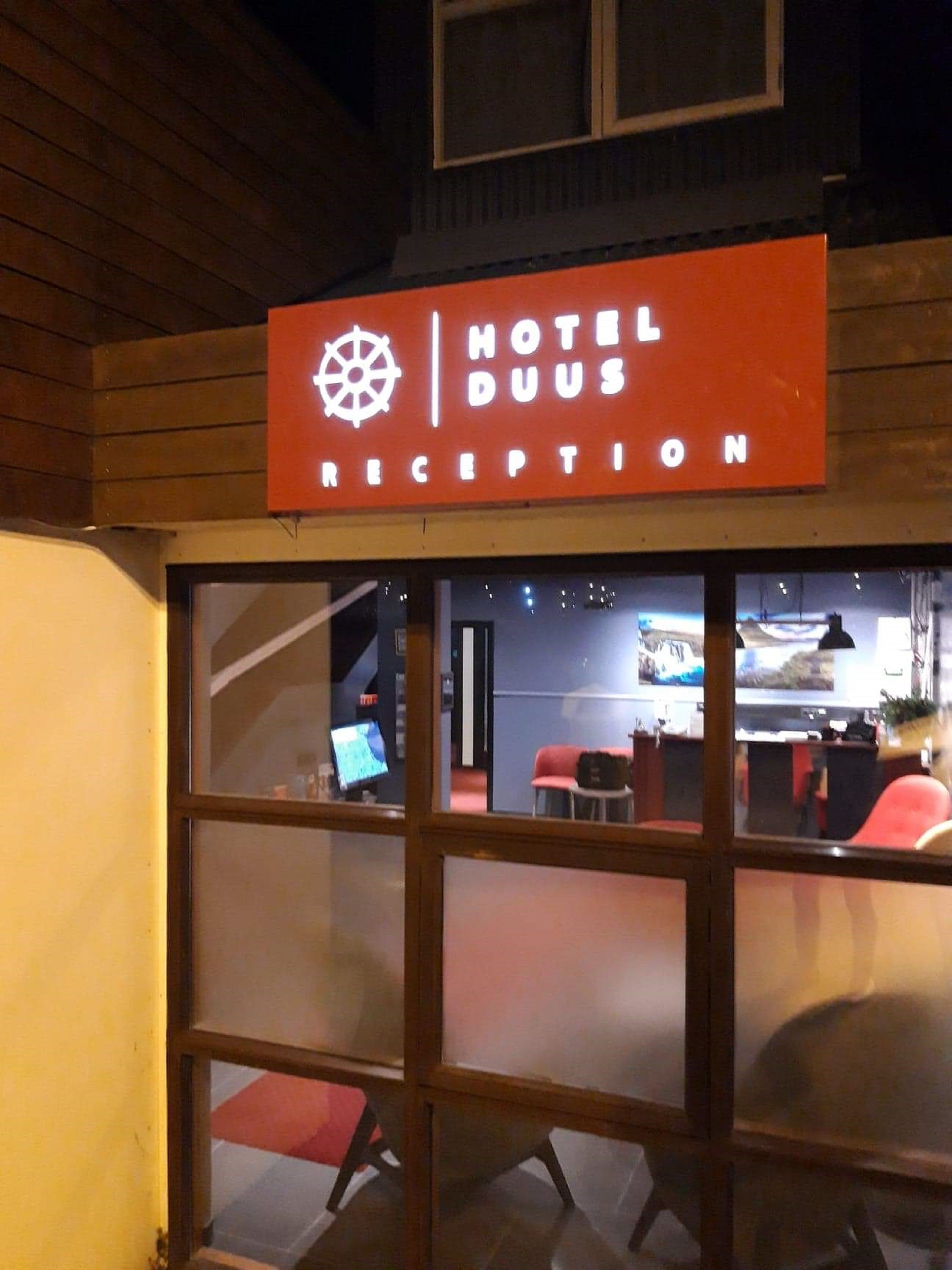 Een hotel receptie met een bordje met de tekst 'HOTEL DUUS RECEPTION' en een zichtbaar bureau met een computerscherm.
