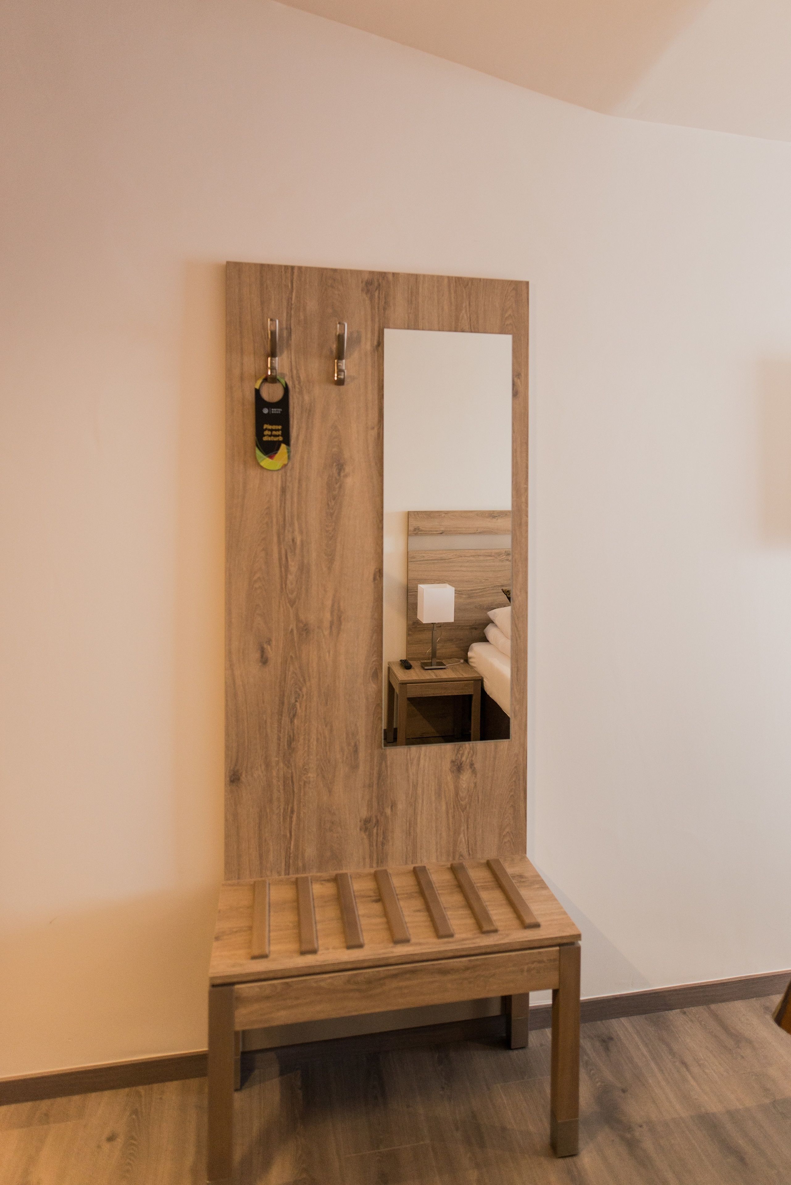 A wooden bench with a mirror attached to a wall, a small bedside table with a lamp, and a wooden floor.