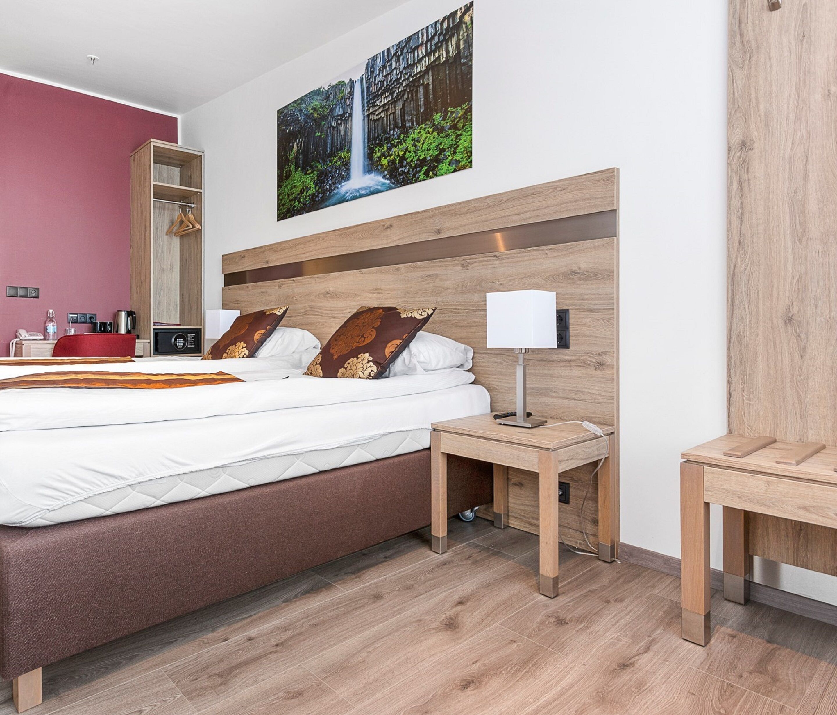 A modern hotel room with a large bed, a bedside table with a lamp, a wooden headboard, and a large mirror.