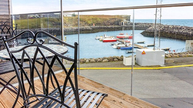 A wooden deck with metal chairs and a table overlooking a harbor with boats and a rocky breakwater. - Hotel Duus (Reykjanesbær)