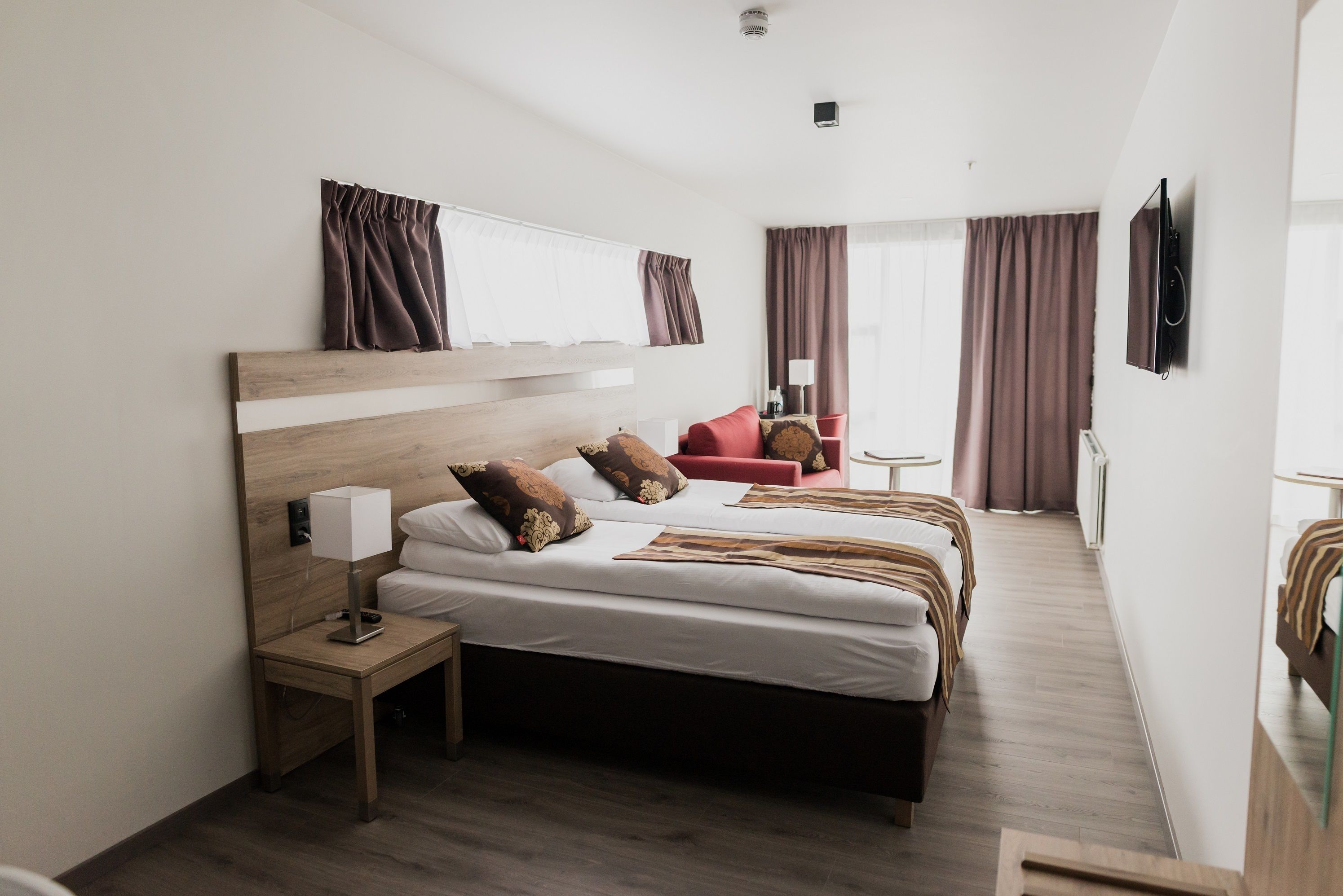 Moderni hotellihuone, jossa on suuri sänky, yöpöytä lampun kanssa, seinälle asennettu televisio ja näkymä huoneen sisustukseen.