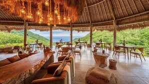 An open-air dining area with wooden furniture and a thatched roof, overlooking a lush landscape.