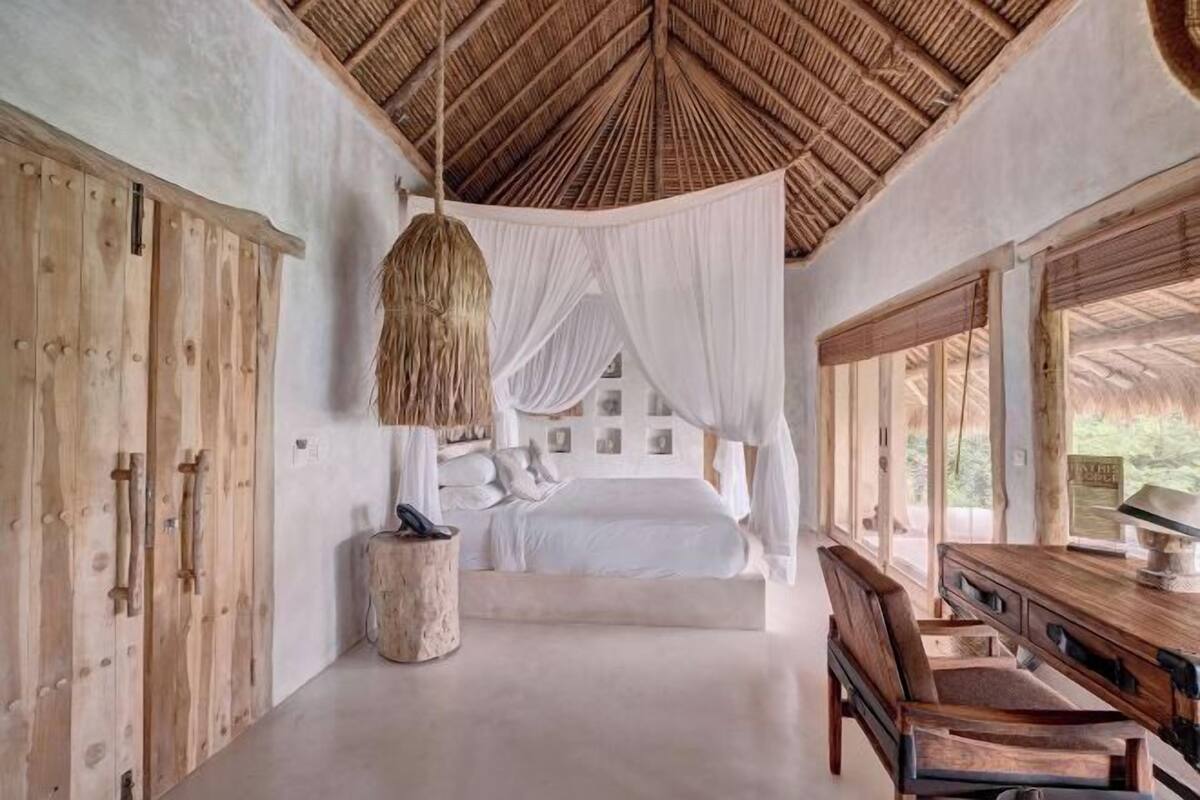 Une chambre avec un toit de chaume, des meubles en bois et un lit recouvert de draps blancs.