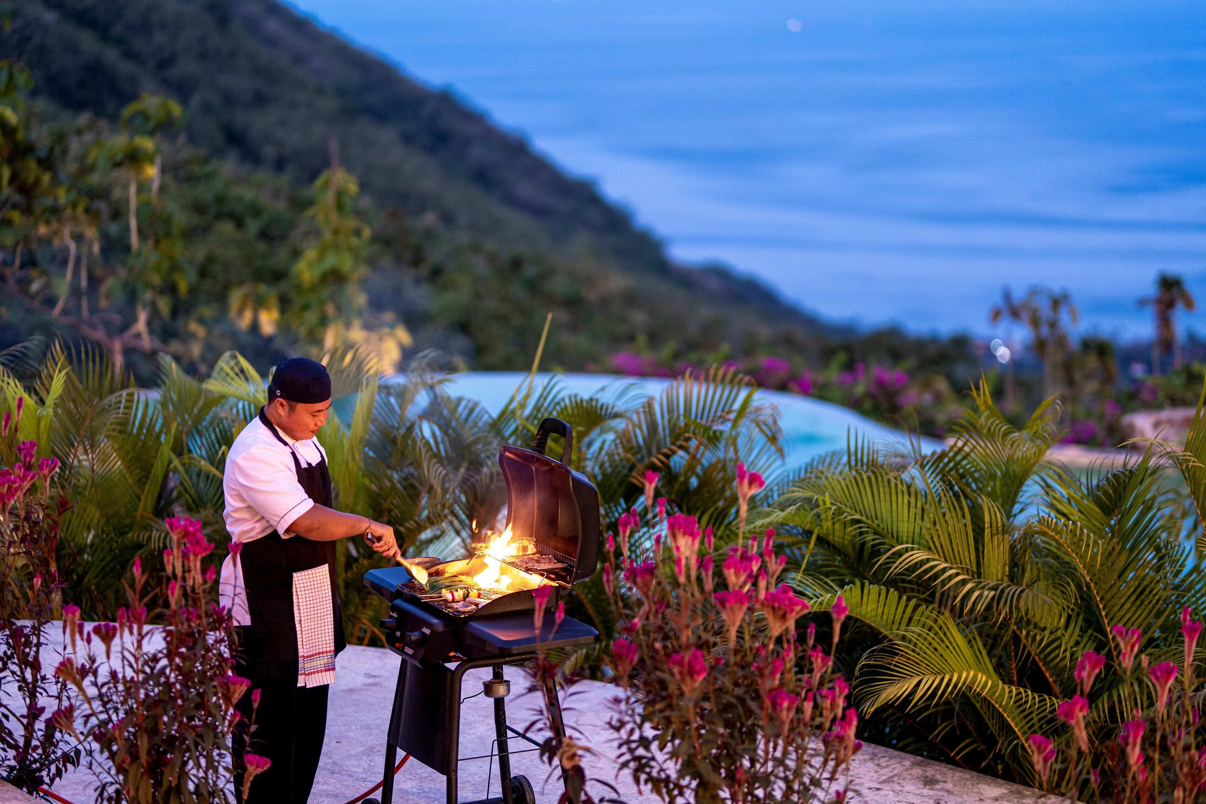 Ein Koch grillt draußen mit Blick auf Strand und Berge im Hintergrund.